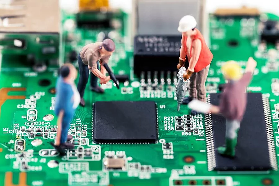 Miniature model figures dressed as construction workers and engineers performing maintenance and repair work on a green printed circuit board.